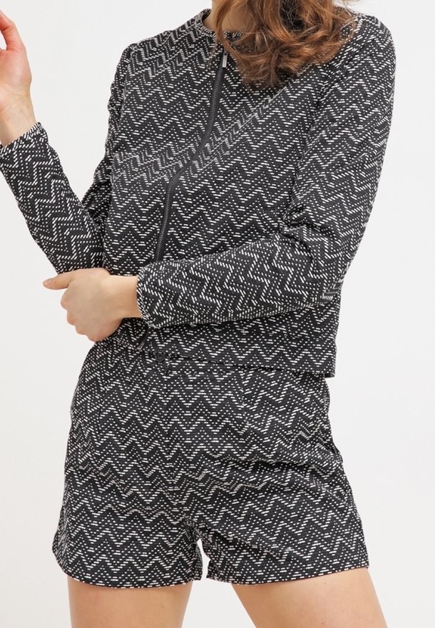 Frau in einer Jacke mit langem Ärmel im schwarz-weißen Zickzackmuster und passenden hochgeschnittenen Shorts, stehend mit verschränkten Armen.