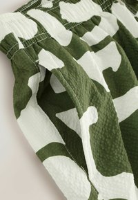 Green and white camouflage-patterned fabric, featuring a textured surface, elastic waist, and gathered detailing on the edges.