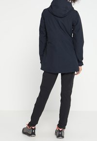 Granatowa wodoodporna kurtka z kapturem, slim fit i elastycznymi mankietami, noszona na czarne spodnie oraz szare buty trekkingowe z różowymi akcentami.