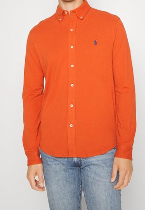 Uomo che indossa una camicia a maniche lunghe di colore arancione brillante con logo ricamato sul petto, abbinata a jeans azzurri.