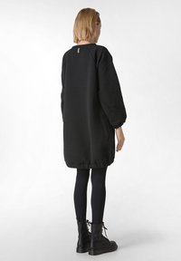 Musta ylisuuri college-mekko, jossa on teksturoitu kangas, raglan-hihat, ribbineuloshelma ja pieni kaulalappu. Pukeutunut mustien leggingien ja saappaiden päälle.
