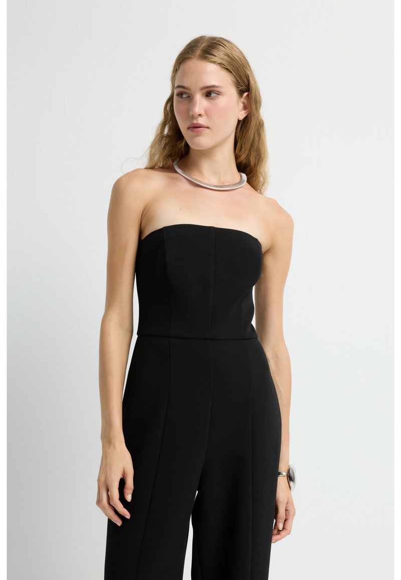 Mono negro sin tirantes con corpiño ajustado y diseño de pierna ancha. Tela suave, costuras mínimas, combinado con un collar de plata.