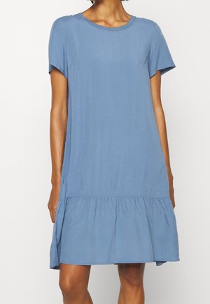 Femme portant une robe bleu clair ample à manches courtes avec un détail froncé au-dessus de l'ourlet qui arrive au niveau du genou.