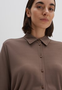 Blouse marron à col, fabriquée en tissu lisse. Elle présente un devant boutonné avec des boutons assortis et une finition texturée.