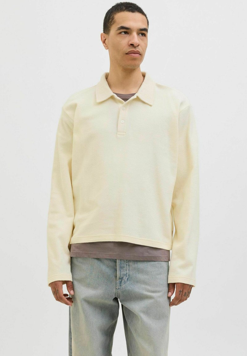 Polo à manches longues jaune clair en tissu texturé, avec un col à boutons, superposé sur une chemise grise, associé à un jean clair.