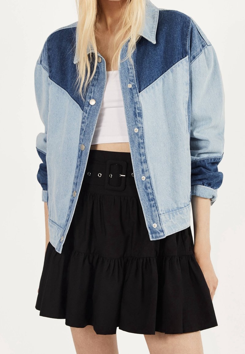 Frau in einer zweifarbigen blauen Jeansjacke über einem weißen Crop-Top und einem hochgeschnittenen schwarzen Rock mit großer Gürtelschnalle.