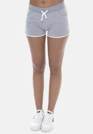 Femme debout portant un short gris avec bordure blanche et des baskets blanches sur fond blanc.