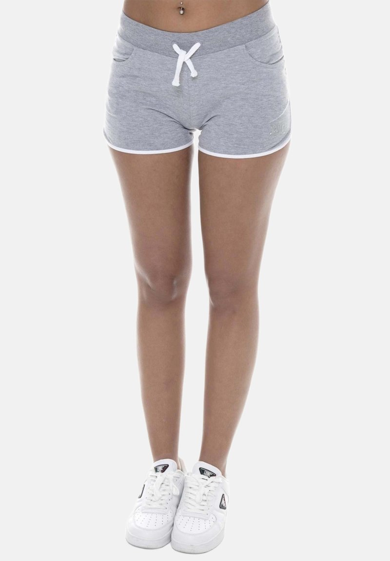 Femme debout portant un short gris avec bordure blanche et des baskets blanches sur fond blanc.