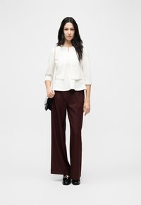 Femme aux longs cheveux foncés portant une veste blanche et un haut blanc, un pantalon large bordeaux, des mocassins noirs, et tenant un petit sac à main avec une bandoulière chaîne.