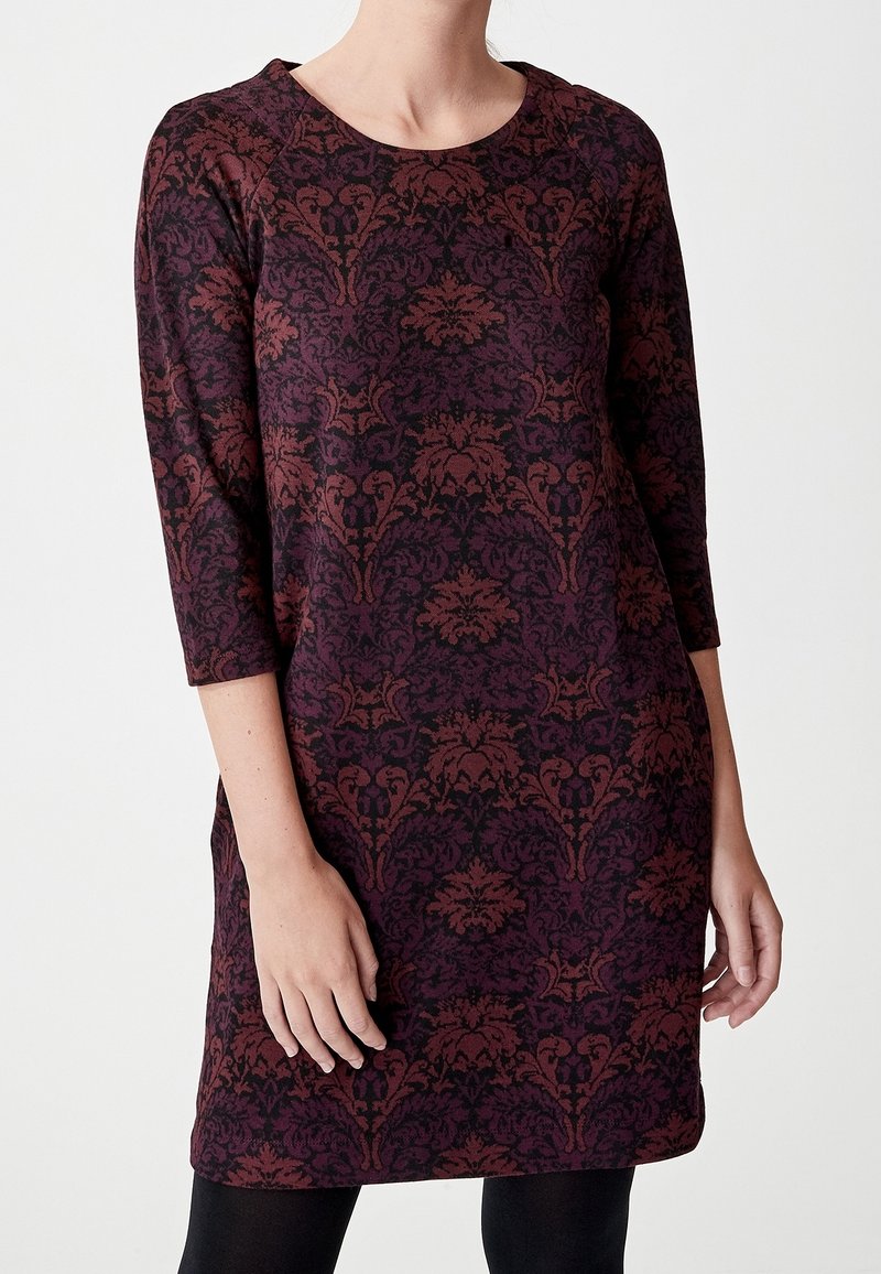 Femme portant une robe à manches longues à motifs violet foncé et bordeaux avec un col rond et des leggings noirs.