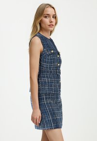 Navyblauw tweed jurk met een getailleerde pasvorm, voorzien van gouden knopen, een vierkante halslijn en een structuurtje. Verstelbare riemdetails.