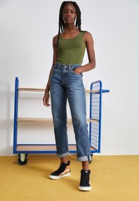 Olijfgroene tanktop, high-waisted blauwe jeans met lichte vervaging, opgerolde manchetten en zwart-tan hoge sneakers. Eenvoudige achtergrond.
