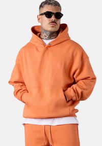 Sweat-shirt à capuche orange en tissu doux, équipé d'une poche avant, de poignets côtelés et d'un logo en relief. Porté avec un jogging assorti.