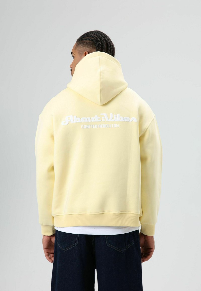 Geel capuchonsweatshirt met de tekst "About Vibes" en "Crafted Rebellion" in wit, geribbelde manchetten en een ontspannen pasvorm.