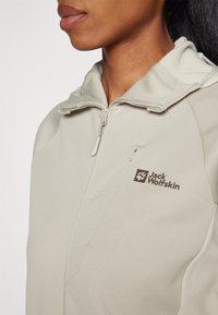 Lichtbeige jas met een ritssluiting aan de voorkant en een capuchon. Heeft een gestructureerde stof en een bruine Jack Wolfskin-logo op de borst.