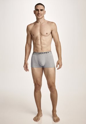 Muscular young man standing barefoot wearing gray Jockey boxer briefs against a plain light background, smiling slightly.