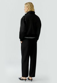 Schwarze Bomberjacke mit cropped Design, mit Stehkragen, getragen über einem grauen Oberteil. Kombiniert mit lockeren schwarzen Hosen und schwarzen High Heels.