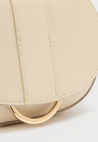 Sac à main en cuir synthétique beige avec une texture lisse, doté d'un accent en métal doré de forme circulaire et de coutures contrastées.