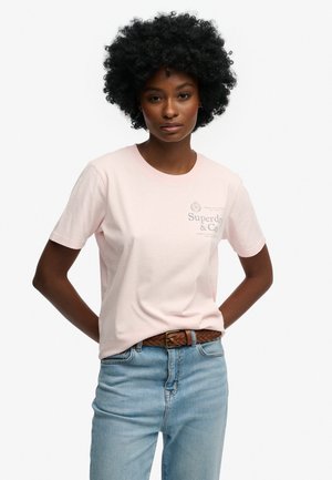 Donna con capelli ricci che indossa una t-shirt rosa chiaro Superdry infilata in jeans azzurri chiari con una cintura intrecciata marrone, in piedi con le mani dietro la schiena.