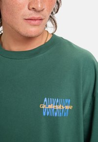 Camiseta de algodón verde con un logo gráfico en azul y naranja. El diseño incluye un cuello redondo y mangas cortas. Tejido texturizado.
