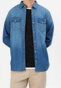 Man som bär en oknäppt blå denimskjorta över en svart t-shirt och beige byxor mot en helt vit bakgrund.