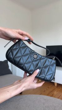 Sort quiltet håndtaske med sølv logo holdt med to hænder i en stue med trægulv og tv i baggrunden.