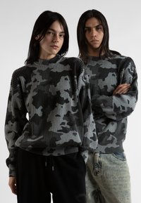 Šedé žebrované svetry s černošedým károvaným vzorem. Modelky nosí volné kalhoty, jedna v černé barvě, druhá ve světlém denimu.