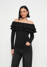 Must ülikõrge kaelaga topp tekstureeritud rüüsidega rinnal, pikad varrukad ja kehakuju rõhutav lõige. Kombineeritud tumedate teksadega.