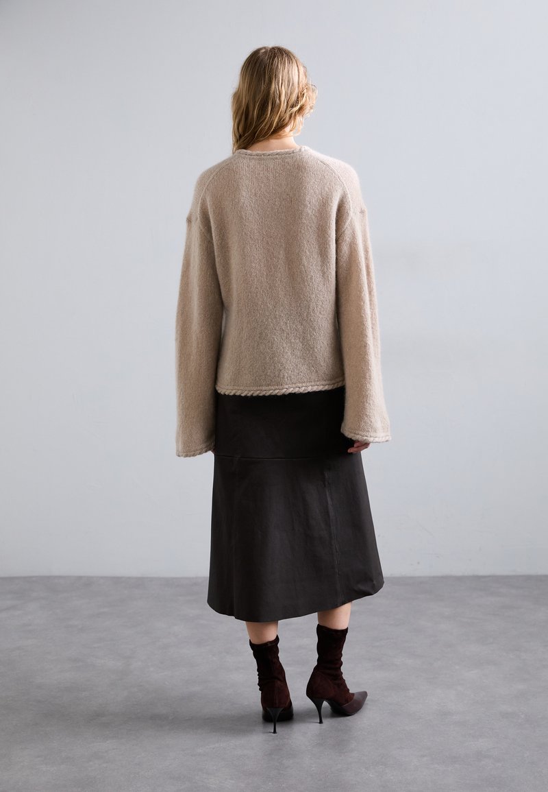 Maglione beige a maglia con spalle scese, abbinato a una gonna midi nera e stivaletti alla caviglia in pelle marrone, presentato su uno sfondo neutro.
