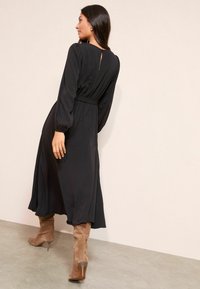 Černé dlouhé šaty s taženým pasem, sklady na zadní části a volným lemem. Nosené s hnědými stiletto botami nad kolena.