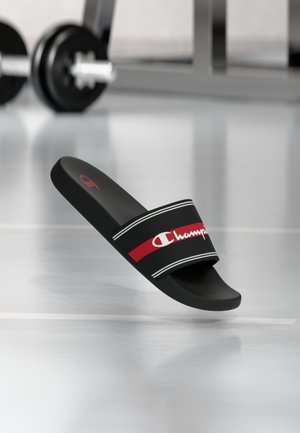 Svarte slip-in-sandaler med bred stropp med en rød stripe og hvite detaljer, med en hvit logo. Flat såle og glatt materiale.