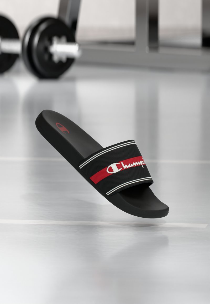 Μαύρη σανδάλι με φαρδιά λουρί που έχει κόκκινη ρίγα και λευκές λεπτομέρειες, με λευκό λογότυπο. Επίπεδο πέλμα και λείο υλικό.