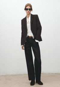 Czarna dopasowana marynarka na białej koszuli, zestawiona z wysokim stanem czarnych spodni. Modelka nosi czarne okulary przeciwsłoneczne i brązowe botki na kostkę.