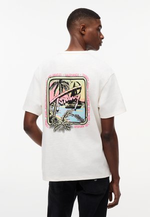 Mann trägt ein weißes T-Shirt mit einem bunten tropischen Sonnenuntergang, Palmen und dem Schriftzug "Tommy" auf der Rückseite, kombiniert mit schwarzen Hosen.