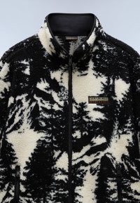 Chaqueta de polar con patrón en blanco y negro, cuello alto, cierre de cremallera y dos bolsillos frontales. Textura suave, con un parche de logo en el pecho.