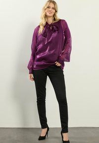 Blusa di raso viola con dettagli a fiocco sul colletto, maniche lunghe trasparenti, abbinata a pantaloni neri slim-fit e tacchi alti neri.