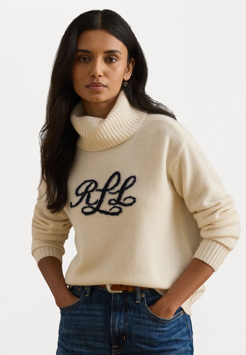 Sweater bege com gola alta canelada, apresentando bordado em azul escuro "RLL" e punhos canelados, emparelhado com jeans de denim azul.