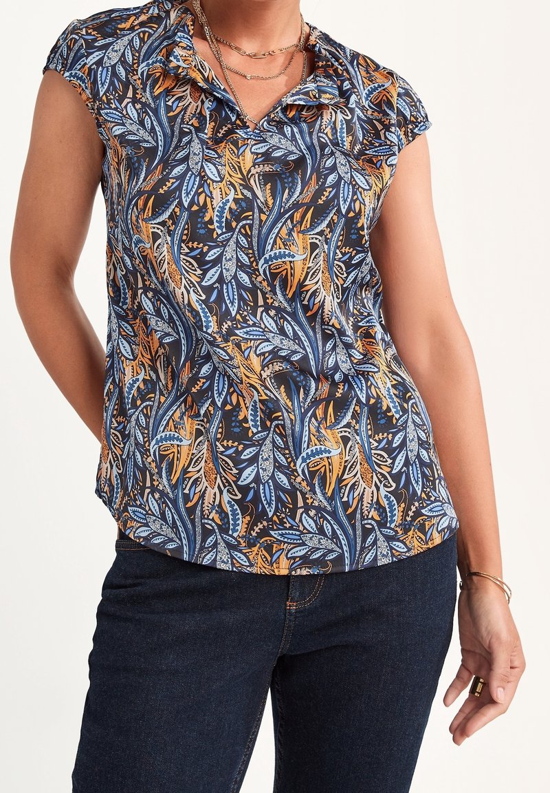 Femme portant un chemisier à manches courtes avec un motif de feuilles bleues et orange, un jean foncé, des colliers superposés et des bracelets au poignet droit.