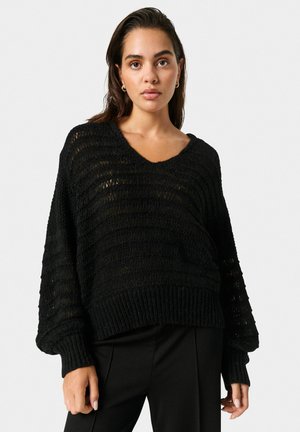 Sort strikket sweater med bred V-udskæring, struktureret design og ribbet kant, parret med skræddersyede sorte bukser.