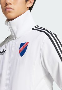 White zip-up jacket with ribbed collar, black stripes on shoulders, and a blue shield logo with "COLO COLO" in red, made of smooth fabric.