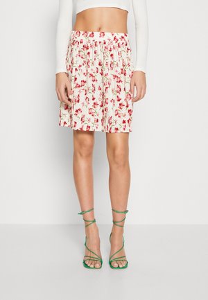 Frau trägt einen weißen Rock mit rotem Blumenmuster, grüne, geschnürte High-Heel-Sandalen und ein weißes, langärmeliges, kurzes Oberteil.