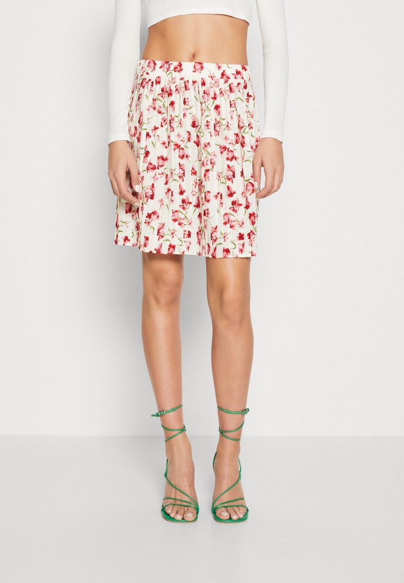 Femme portant une jupe blanche à imprimé floral rouge, des sandales vertes à talons hauts avec des lanières, et un haut court blanc à manches longues.