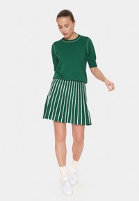 Haut tricot vert à manches courtes, associé à une jupe plissée verte avec des rayures verticales blanches, et des baskets blanches avec des chaussettes légères.