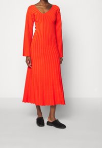 Robe rouge côtelée à manches longues et col en V, avec un ourlet évasé. Associée à des chaussures noires sans lacets, sur un fond neutre.