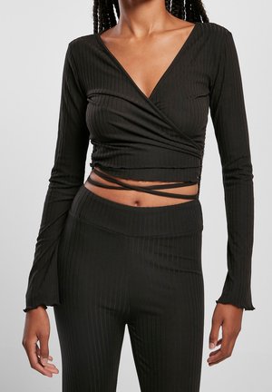 Femme portant un crop top cache-cœur noir côtelé à manches longues avec liens et un pantalon côtelé taille haute, sur un fond gris uni.