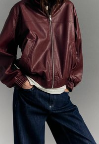 Burgundowa skórzana kurtka bomberka o skróconym fasonie, z zapięciem na zamek, ściągaczami przy mankietach i bocznymi kieszeniami, zestawiona z ciemnoniebieskimi szerokimi spodniami dżinsowymi.