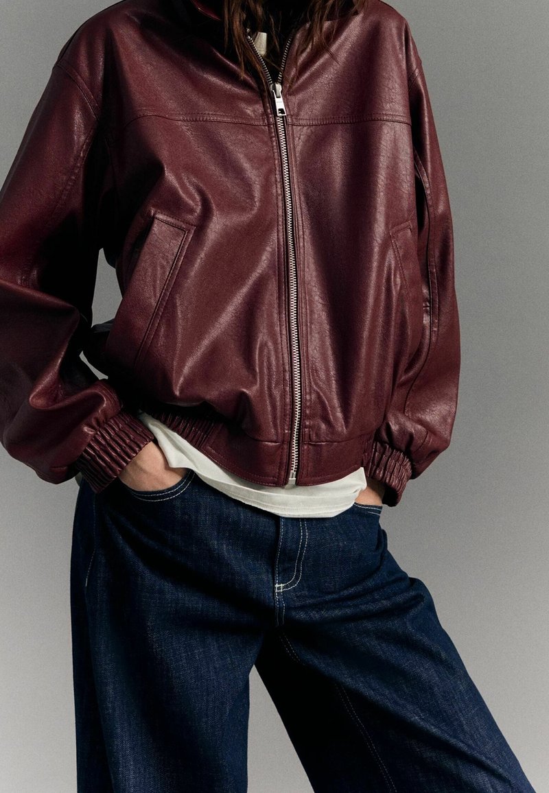Burgundowa skórzana kurtka bomberka o skróconym fasonie, z zapięciem na zamek, ściągaczami przy mankietach i bocznymi kieszeniami, zestawiona z ciemnoniebieskimi szerokimi spodniami dżinsowymi.