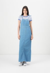 Abito lungo in denim con spalline regolabili, tasca frontale, taglio dritto, lavaggio azzurro chiaro, abbinato a sneaker grigie e una maglietta a righe.