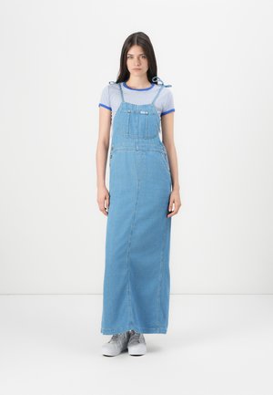 Denim maxi-jurk met verstelbare schouderbanden, voorzak, rechte snit, lichtblauwe wassing, gecombineerd met grijze sneakers en gestreept T-shirt.