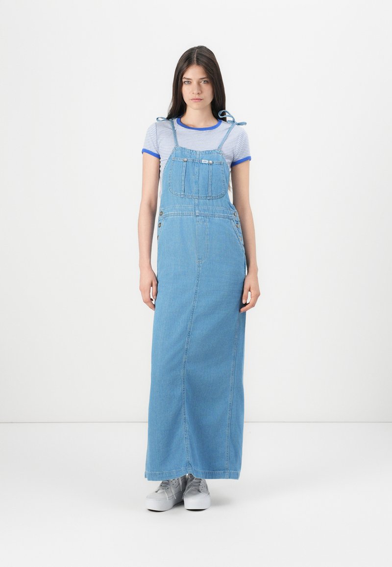 Abito lungo in denim con spalline regolabili, tasca frontale, taglio dritto, lavaggio azzurro chiaro, abbinato a sneaker grigie e una maglietta a righe.